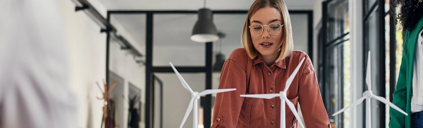 Une femme aux cheveux blonds raides, portant des lunettes et une chemise orange à manches longues, regarde un modèle de trois petites éoliennes.