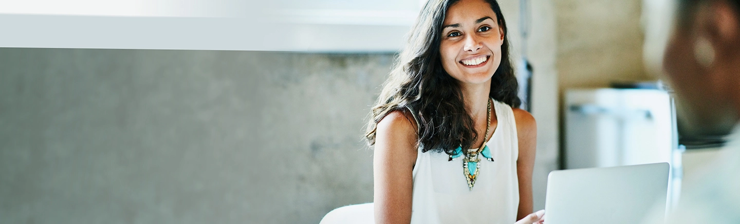 Brunette with teal necklace smiling at diverse colleague in office setting