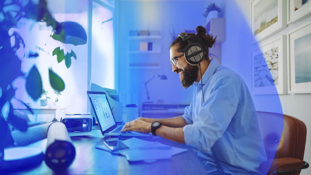 A man on his laptop smiles as her works in his home office. He is hearing headphones and a blue dress shirt.
