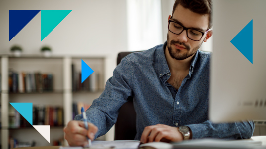 Un homme en chemise bleue et lunettes prend des notes sur son bureau avec un ordinateur à sa droite.