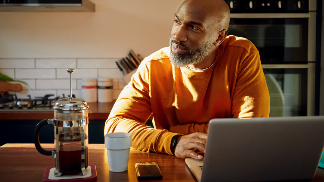A man on a bright orange sweater sits on his kitchen with a cup of coffee and his laptop in front of him.