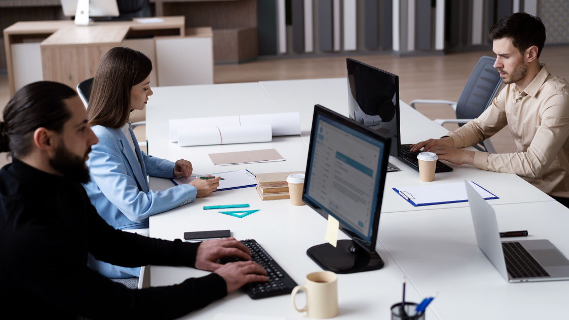 A group of two men and a woman work on a desk in an office. The men work on monitors while the woman writes on a notepad. Around them there are coffee cups, a laptop, papers and another notepad.