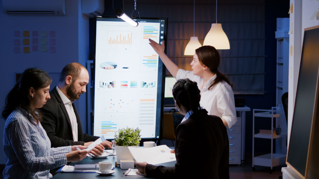 A group of people look at data and analytics in a vertical monitor inside a darkened office. 