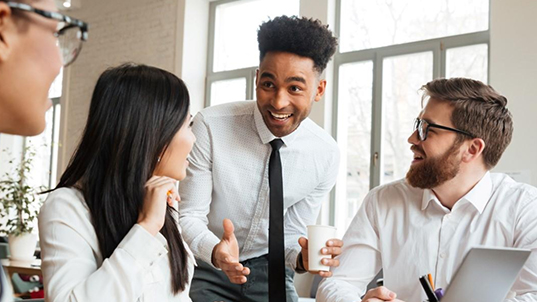 Two men and a woman chat enthusiastically on an office setting. One fo them has a coffee cup.