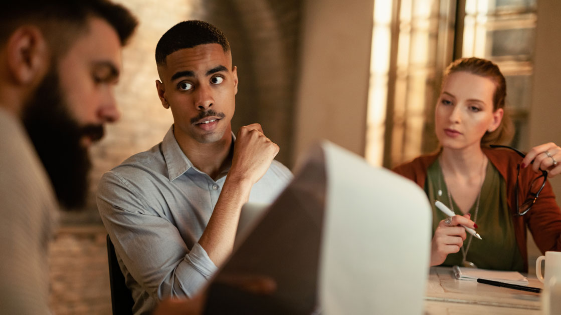 A man and a woman look with interest at another man as he shows them something on his clipboard. They're all in an office as sunset light filters through.