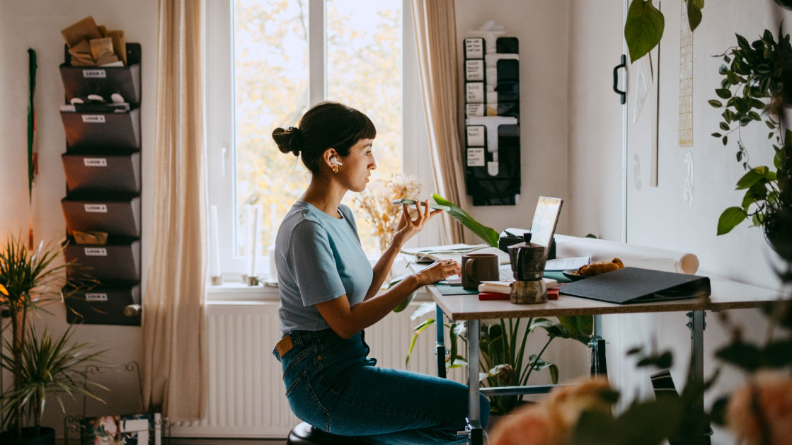 A woman works from home, using her laptop, earphones and talking into her cellphone. She is in a white living room surrounded by plants with a pot and cup of coffee.