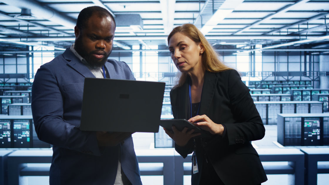A man and a woman look at a laptop inside a server room.
