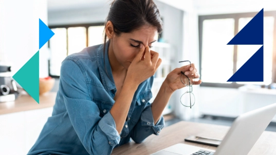 A woman wearing a blue shirt sits in front of her laptop. In one hand she holds her glasses and with the other she squeezes the bridge of her nose in frustration. 