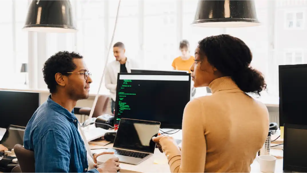 Deux personnes discutent dans un bureau avec un ordinateur affichant du code en arrière-plan.