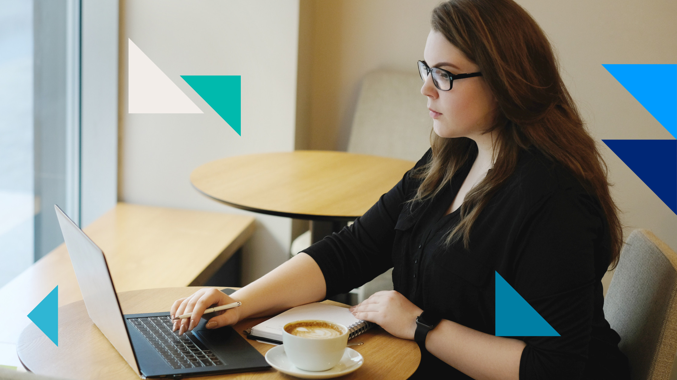 A woman works in a cafeteria. In front of her on a desk is a laptop, a notebook and a cup of coffee.