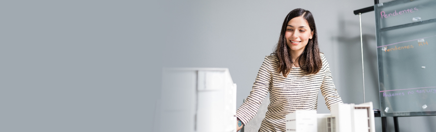 A woman with a beige striped shirt overlooks some model buildings with a smile. Next to her there's a transparent whiteboard with some notes in different colors.