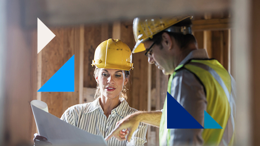 Close up of a man and woman wearing yellow hard hats looking on and discuss a plan blueprint on a construction site.