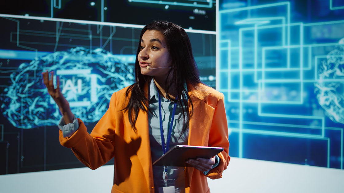 Une femme vêtue d'une veste de tailleur orange vif parle dans ce qui ressemble à une présentation avec une image lumineuse d'un cerveau et un panneau indiquant AI.
