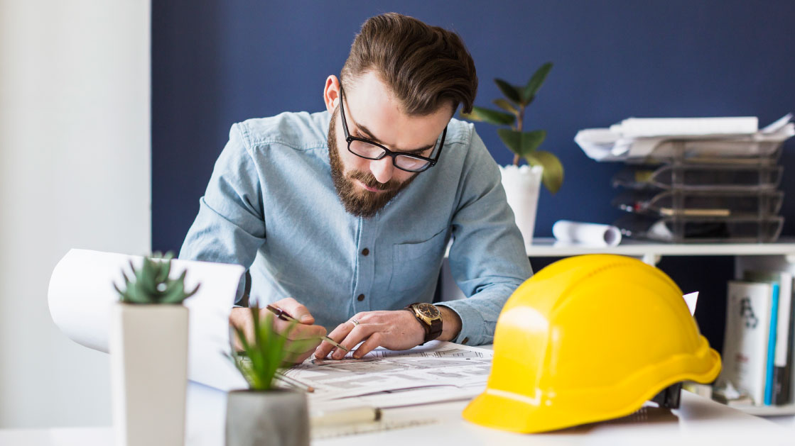 A man looks at plans while sitting on a desk. In front of him is a yellow construction hard hat.