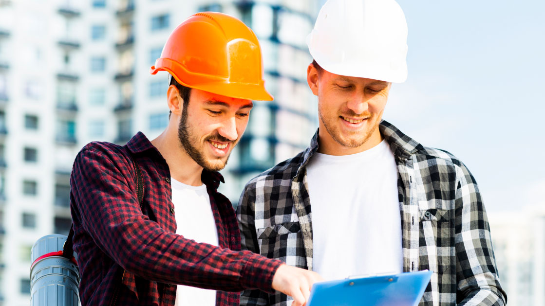 Two men in plaid shirts and construction hard hats look at a notepad. There's a blurry apartment building behind them.