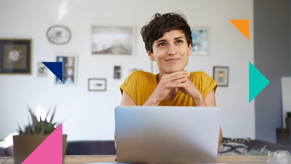 A woman with a mustard-colored t-shirt looks happy with a small smile on her face. There's a laptop in front of her and multiple frames with pictures hanging on a white wall on the background.