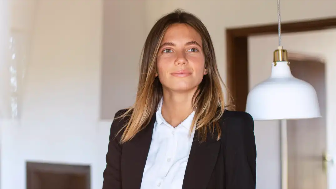 Une femme vêtue d'une chemise blanche et d'une veste de costume sombre sourit dans un bureau.