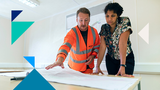 A man in bright orange construction gear shows a construction plan on a table to a woman wearing a black and white blouse.