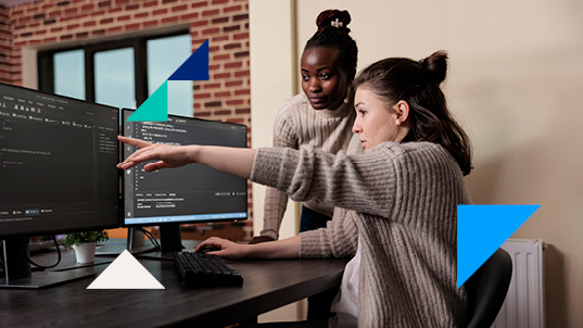 Two women look at double monitors displaying a coding program on an office. One of the women is pointing at something on the screen.