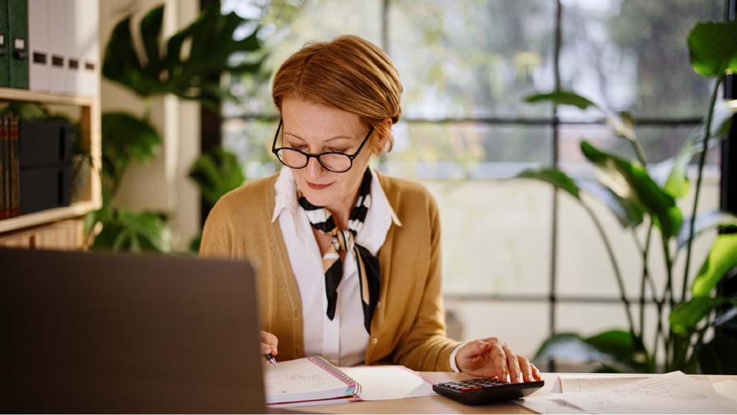 A woman works from home on her living room. She is writing something on her notepad and using a calculator. Behind her are plants and a large window.