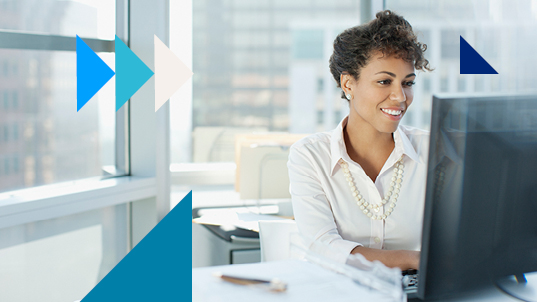 A woman wearing a white blouse and a pearl necklace smiles as she works in an office with big windows.