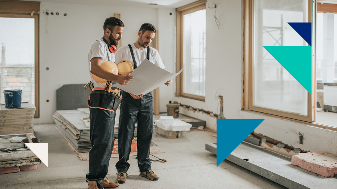 Two men wearing construction gear stand in a construction site and look at a blueprint.