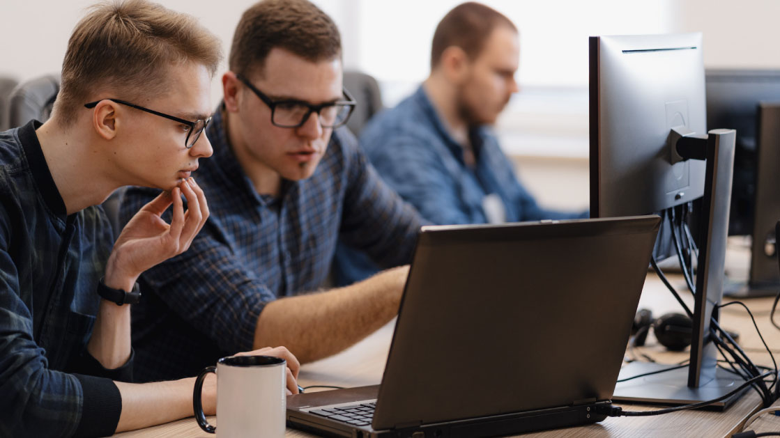 Two men wearing black glasses and blue plaid shirts work on a laptop and a monitor with interest. Behind them is a third man, also on a blue plaid shirt, working on his own monitor.