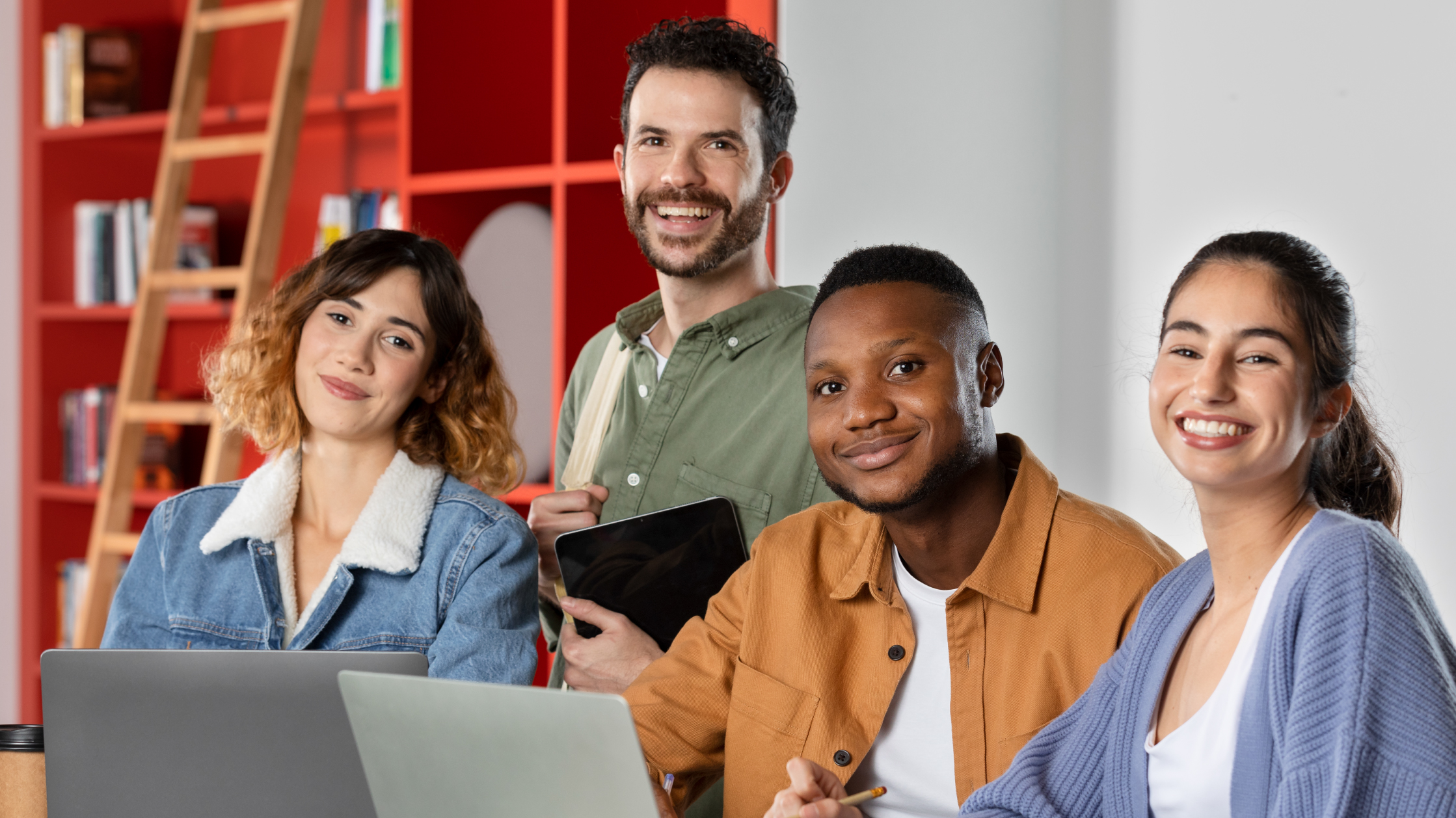 A group photo of young women and men wearing colorful clothes with laptops and tablets. Behind them is a bright red bookshelf with a stair.
