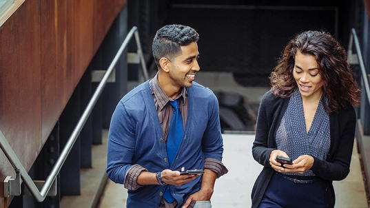 A man and a woman walk up some stairs with their cellphones on their hands. The man is looking at the woman and smiling.