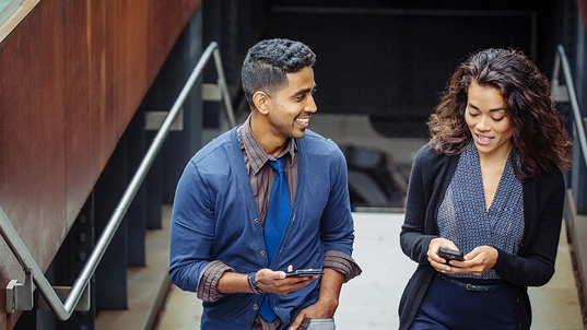 Un homme et une femme montent des escaliers en tenant leur téléphone portable à la main. L'homme regarde la femme en souriant. 