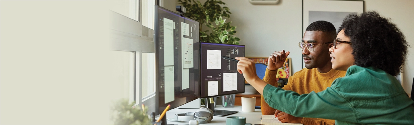 A man looks attentively at two computer monitors in front of him as a woman points with a pen.