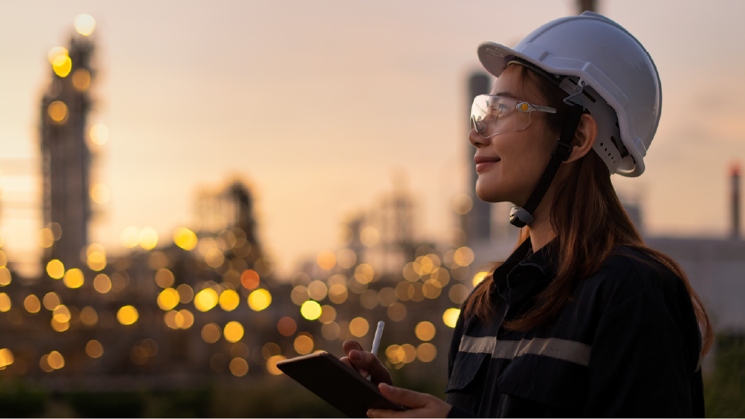 A woman in protective gear, a white helmet, plotective goggles and black vest, makes some annotations as she looks on. Behind her, unfocused, are bright yellow lights of a city.