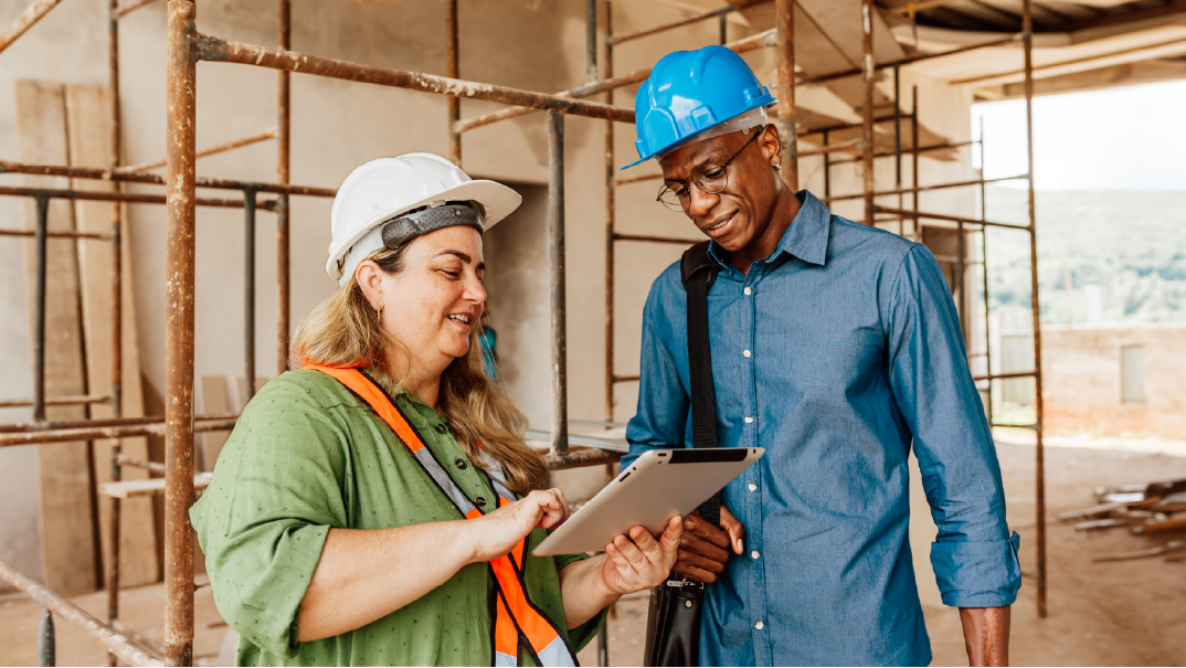 A man and a woman stand in a construction site and look at a tablet on the woman's hands. They are both wearing safety hardhats, white and blue.