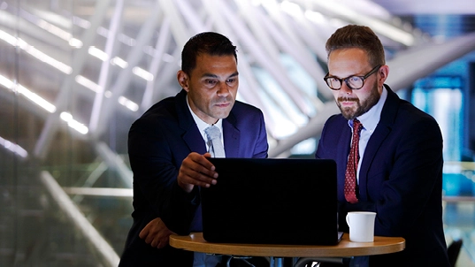 Two men wearing navy blue suits look into a compuer. One of the wears a red tie and glasses and the other a grayish-blue tie.