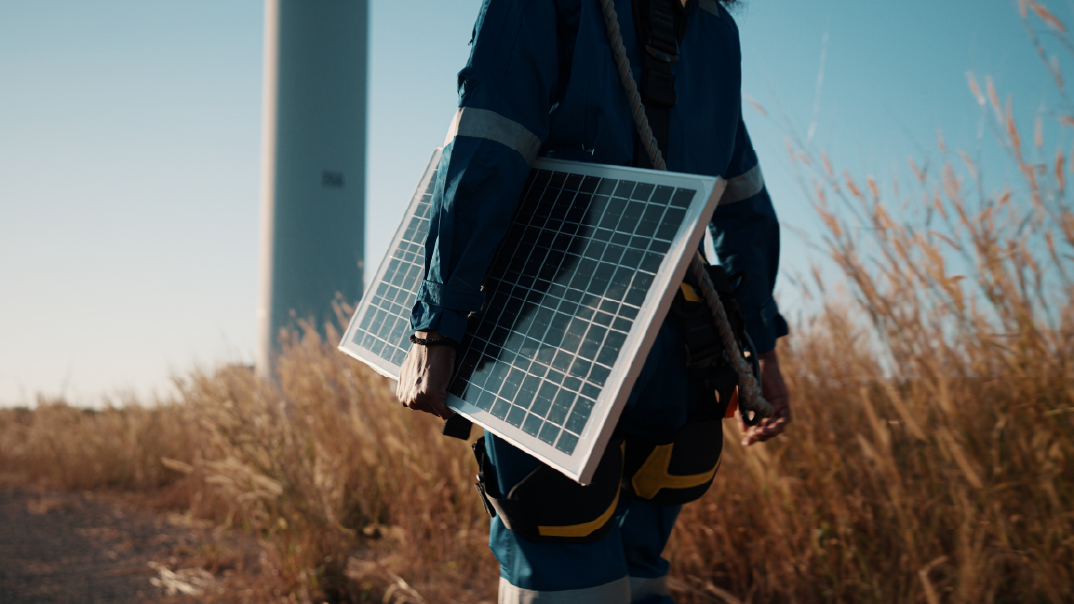 A man walks on a field of grass carrying a solar panel.