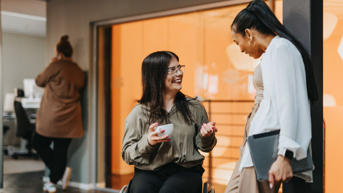Two women in conversation indoors, one holding a mug and seated, the other standing with a tablet. A third person is in the background.