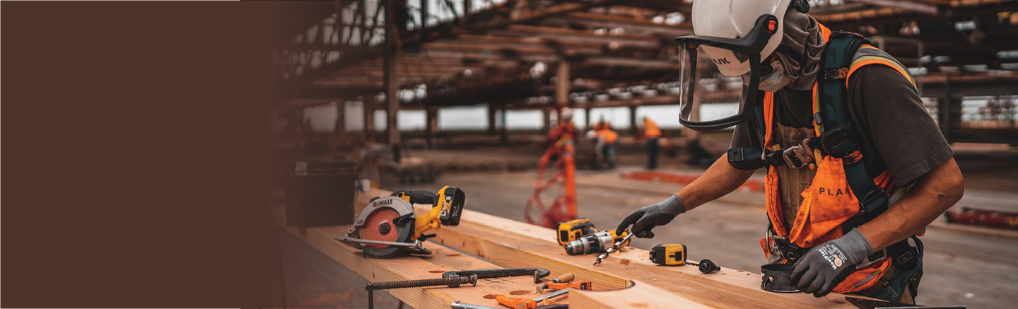 A man in safety gear including a white hardhat works on a piece of wood in front of him. There are several yellow and orange tools on it.